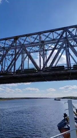 The camera looks upwards at a grey railway bridge spanning a wide blue river. The bridge's frame is comprised of interlocking steel beams. A train, consisting of dark, possibly green, railcars, is seen travelling across the bridge. The river stretches to the horizon beneath a blue sky dotted with white clouds. A small boat is seen in the distance on the river. The camera is positioned on a boat or ship. A part of the ship's equipment is visible in the lower right corner of the frame. Some trees can be seen on the riverbanks.