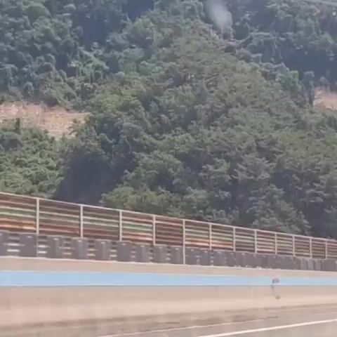 A first-person perspective video shows a highway drive. The camera is facing forward, capturing the road and the surrounding environment. To the left, a lush green mountainside covered in dense trees rises steeply. A tall, grey pole, likely a lamppost, stands prominently in the center of the frame against the mountainside. A noise barrier, constructed of horizontal wooden planks and metal supports, runs along the right side of the highway. The road has multiple lanes, indicated by white and blue lines. In the distance, a red bus with "VIP" written on its back is visible, along with other vehicles. The sky is partly cloudy, with mountains visible in the far distance. The video captures the motion of the vehicle as it travels along the highway, with trees, mountains, and the noise barrier blurring slightly due to the speed.