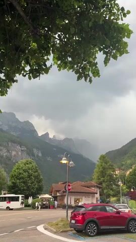 The video shows a scenic view of a mountain town with a dramatic cloudy sky. In the foreground, a red car is parked in a parking lot. A street light is visible behind the car. Further back, there is a building with outdoor seating and people walking around. A bus is parked on the left side of the frame. The background features lush green mountains under a dark, overcast sky, with a hint of rain or mist in the distance. The overall atmosphere is one of impending weather change in a picturesque European setting.