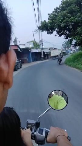 A first-person perspective video shows a person riding a motorcycle on a paved road. The rider's hands are visible on the handlebars, and a child's head is visible in the foreground. A motorcycle mirror reflects a person wearing a bright green shirt. The road curves to the right, with shops and buildings lining the left side of the road. A truck is visible in the distance. The sky is overcast. The video appears to be taken during daylight hours.