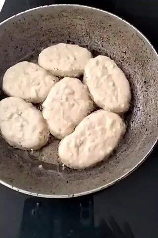 The video shows a close-up view of several oval-shaped food items being fried in a pan. The items are pale in color and appear to be some kind of fritters or pancakes, possibly made from a batter with small lumps. The pan contains oil, and some of the items show slight browning at the edges. The camera then pans out to show the pan on a stovetop and then shifts to a plate of the same food items, now golden brown and cooked, resting on a white paper towel. A fork is visible next to the plate. The background suggests a kitchen or dining area.