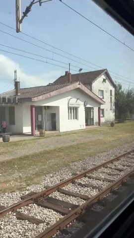 The video shows a view from a moving train, passing by a small train station building labeled "GOLUBOVCI ГОЛУБОВЦИ". The station is a white, two-story building with a small porch supported by magenta columns. There is a grassy area surrounding the building with train tracks and ballast stone in the foreground. A man wearing a dark uniform and hat walks in front of the station building. Overhead, electrical wires are visible against a partly cloudy sky.