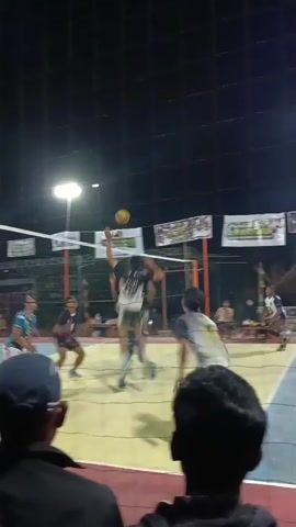 A volleyball match is underway at night. Several male players are actively engaged on a court, with a net dividing the two sides. In the foreground, two individuals, presumably spectators, are partially visible from behind, their heads and shoulders in view. The court is illuminated by overhead lights, and the background reveals a dark, presumably outdoor, setting with a netted enclosure. The players are wearing athletic attire, and the game appears to be in progress, with some players in mid-action.
