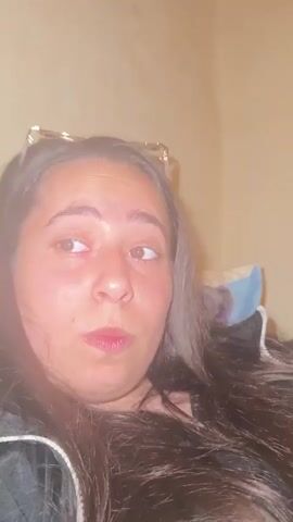 The video shows a close-up of a young woman's face as she appears to be experiencing various emotions. She has dark hair and is wearing glasses on top of her head. Her expression shifts from what seems like surprise or confusion to a more neutral look, and then back to a surprised or shocked expression with her mouth open. The background is a plain, light-colored wall, and some blurred objects, possibly pillows or blankets, are visible behind her. The lighting suggests an indoor setting, possibly at night or in a dimly lit room.