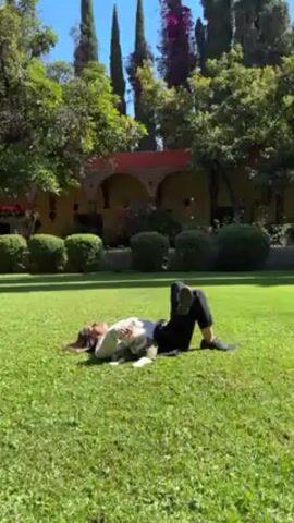 A woman is lying on her back on a lush green lawn, with her legs bent at the knees and her feet flat on the ground. She is wearing a light-colored sweater, dark pants, and dark shoes. A small dog is lying on her chest. The dog is wearing a vest and is light in color with patches of dark fur. The woman appears to be petting the dog. The setting is a garden with trees, bushes, and a building in the background. The building has arches and a red tile roof. The weather is sunny and bright.