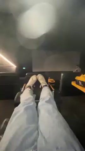 A first-person perspective video shows a person reclining in a movie theater seat, their legs stretched out towards the screen. The person wears jeans and light-colored shoes, with colorful socks peeking out. A tray table next to them holds a large container of yellow food, likely popcorn or fries. The camera then turns to reveal a young woman with long dark hair with red highlights, wearing a gray sweater over a black top. She smiles at the camera. The environment is dimly lit, consistent with a movie theater.
