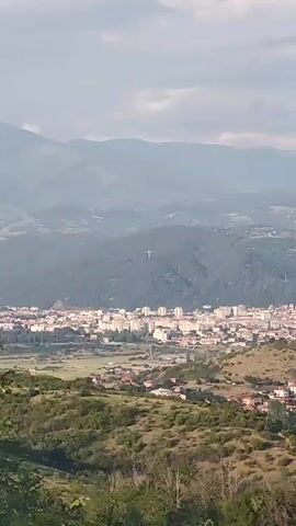 The video presents a panoramic daytime view of a town in a valley, nestled amidst rolling green hills and distant mountains, likely in the region of Selishte, Bulgaria. The sky is partly cloudy or overcast, casting a soft, somewhat hazy light over the landscape.

The primary subject of focus is a large, white cross standing prominently on a forested mountainside in the distance, overlooking the town. The camera slowly zooms in on this distinctive landmark.

The town itself features numerous buildings, predominantly multi-story structures with light-colored facades, suggesting residential and possibly commercial areas. Roads and infrastructure are visible within the town layout. In the foreground, below the vantage point, are more green, shrub-covered hills.

No people are visible. There is no discernible text in the video. The scene evokes a sense of tranquility and grandeur, emphasizing the natural beauty of the mountainous terrain and the striking man-made landmark of the cross.