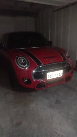 A red Mini Cooper with black racing stripes is parked inside a garage. The license plate reads "PDR 4612". The garage floor is grey concrete. The lighting is dim.