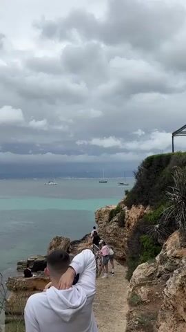 The video shows a coastal view on an overcast day. The foreground features a rocky path, with people walking along it. A man in a white hoodie stands in the foreground with his back to the camera. The middle ground reveals a body of water, with several sailboats visible on the horizon. The sky is filled with heavy, grey clouds, suggesting potential rain. The scene has a natural, somewhat somber atmosphere.