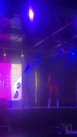 The video shows a stage performance with two people. The stage is lit with vibrant purple and blue lights. On the left side of the stage, there is a large screen displaying a pink graphic with white text and shapes. To the right of the screen, a person stands on stage holding a microphone. Further to the right, another person is also on stage, also holding a microphone and standing near a small table with bottles on it. The second person appears to be sitting on a stool. The lighting creates silhouettes of the performers. The overall atmosphere is that of a live music or performance event.