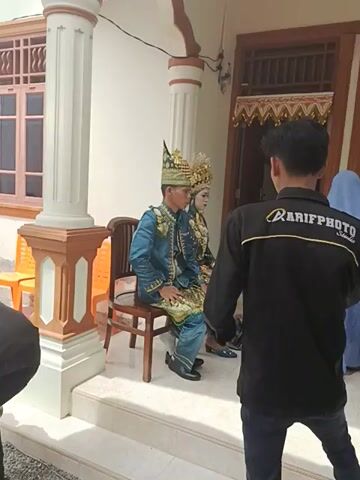 Two people dressed in traditional formal wear are sitting on chairs. One person is taking their picture. All three people are indoors, standing in front of a house with a white porch. The person taking the picture is wearing a black jacket that says "ARIFPHOTO Studio".