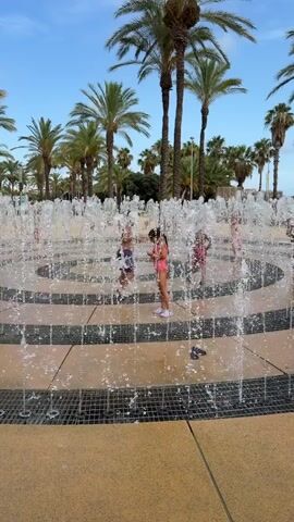 The video shows an outdoor water fountain feature with multiple concentric rings of spraying water. Several children are playing within the fountain. The ground around the fountain is paved with circular sections of tan and dark gray tiles. In the background, there are tall palm trees and a clear blue sky.