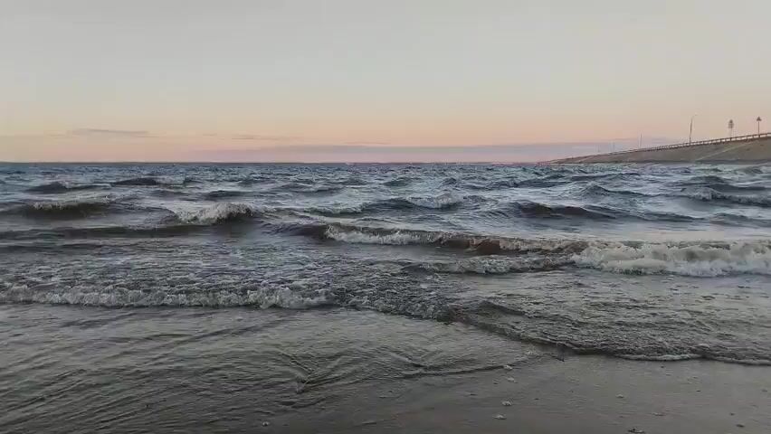 The video shows a close-up, low-angle shot of waves rolling onto a sandy beach at dusk. The water is a dark, choppy grey with white foam cresting the waves. The sky above transitions from a pale pink at the horizon to a clear, light grey higher up. A distant, dark landmass is visible on the horizon to the left, and a concrete structure, possibly a bridge or dam, is visible on the right horizon. The sand in the foreground is dark and wet, with small debris scattered on it. The camera angle remains static, focusing on the movement of the water and the interplay of light on the waves and sand.