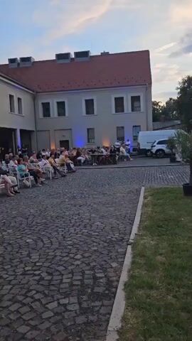 An outdoor event is taking place in a cobblestone courtyard with a light-colored, two-story building in the background. The building has a red-tiled roof and a series of windows and entrances along the ground floor. A group of people are seated in white chairs arranged in rows facing the building, suggesting a performance or presentation. Another group of people are standing and mingling in the covered walkways of the building. The time of day appears to be dusk, with soft lighting from various sources illuminating the area. A white van is parked to the right of the frame, near the building. The overall atmosphere suggests a cultural or social gathering.