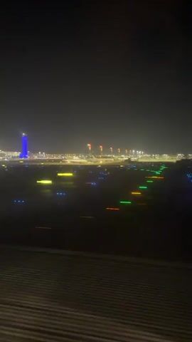 A nighttime view from an airplane window shows an airport runway at dusk or night. Lights from the runway and other airport facilities are visible. In the distance, a tall tower with blue lights illuminates the sky. The image is slightly blurred, suggesting movement.