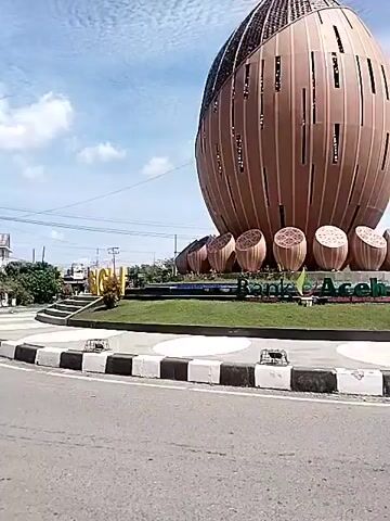 The video shows a large, brown, ovular monument in the middle of a roundabout. The monument has vertical slits or panels running along its side. Around the base of the monument are several smaller, round, brown structures that appear to be decorative. There is a green lawn around the base of the monument. In front of the monument, the word "SIGI" is spelled out in gold letters on the lawn. The roundabout has a black and white striped curb. The road around the roundabout is gray asphalt. There are power lines, buildings, and trees visible in the background. The sky is blue with some white clouds. The video is taken from inside a moving vehicle.