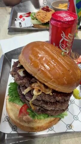The video shows a close-up view of a meal on a table. In the foreground, a large burger with multiple patties, lettuce, tomato, and fried onions is featured prominently. To the right of the burger is a can of Coca-Cola. Behind the burger, a tray holds a chicken sandwich with lettuce and tomato, another can of Coca-Cola, and a bowl of french fries. The scene appears to be in a restaurant or food court, with other trays and a paper placemat visible. The lighting is bright, suggesting an indoor setting.