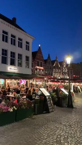 This video captures a lively evening scene in Bruges, Belgium. The setting is a cobblestone square lined with historic buildings, many of which feature ornate facades with red-tiled roofs and tall windows. The time of day is dusk or early evening, as indicated by the deep blue sky and the warm glow of streetlights and illuminated restaurant patios.

The square is bustling with people. In the foreground, a few individuals walk across the cobblestones. Further back, numerous people are seated at outdoor tables of restaurants and cafes, enjoying meals and drinks. The atmosphere appears convivial and relaxed. Plants in green planters add touches of color to the scene.

Notable landmarks are visible. A tall, ornate bell tower, likely the Belfry of Bruges, stands prominently in the distance. Several other grand buildings with impressive architecture are also present. The presence of bicycles parked in front of some establishments suggests a common mode of transport in the area.

The activity is primarily centered around dining and socializing outdoors, taking advantage of the pleasant evening ambiance. People are seen walking, talking, and enjoying the city's atmosphere. The camera work is somewhat dynamic, with some slight motion blur adding to the sense of movement and activity.

Visible text includes a sign on one of the buildings that appears to read "Le Salles" or a similar name, suggesting a specific establishment. A yellow and blue flag, possibly the flag of Ukraine, is also visible, adding a layer of international context. The overall mood conveyed is one of a charming European city coming alive in the evening.
