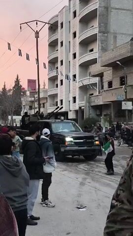The video shows a group of people standing on a street as a black pickup truck drives by. The truck has mounted artillery in the back, and there are people riding on the back of the truck as well. The people on the street are watching the truck as it drives by, and some are taking pictures or videos with their phones. The street is lined with buildings, and there are decorations hanging between the buildings. There is a sign in the distance that says "ROSE". Several children run in front of the truck. The time of day appears to be dusk, as the sky is pink.