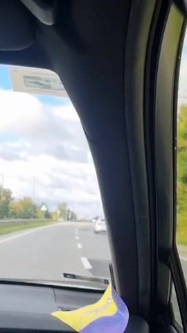 The video shows a first-person view from inside a car traveling on a highway. The camera is positioned looking forward through the windshield and partially through the passenger side window. The road ahead is visible, with multiple lanes and other vehicles in the distance. Trees and power lines line the sides of the highway, and the sky is partly cloudy. A car air freshener is attached to the dashboard in the foreground. The video is short and shows continuous movement, indicating the car is in motion.