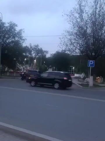 The video shows a street scene with several cars parked or driving. The sky is overcast, and it appears to be late afternoon or early evening. There are trees and buildings visible in the background. A blue parking sign is also visible on the right side of the screen. The camera is positioned on the street, looking towards the parked cars and buildings. The video is short and does not show any significant events or actions.