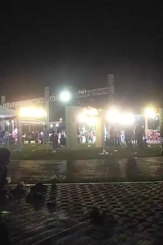The video shows a nighttime outdoor event. In the foreground, there is a paved area with some loose items scattered on it, and in the background, there is a grassy area with people gathered. Large illuminated structures, likely stages or lighting rigs, are visible. There are bright lights illuminating the area, and the ground appears wet, suggesting recent rain. People are seen standing and moving around the illuminated structures.