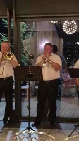 A man in a white shirt and black pants plays a trumpet in front of a glass wall. He is part of a band, as another man to his left plays a similar instrument, and a man to his right plays a saxophone. Music stands with sheet music are placed in front of each musician. The background shows people sitting at tables and a decorative light fixture.