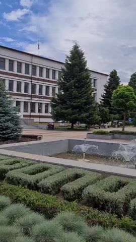 A video displays a public square or park scene. In the foreground, a rectangular water feature contains multiple fountains spouting water. Below the fountains, hedges are shaped to spell the word "LOVE." To the left, a multi-story building with numerous windows stands. As the camera pans right, another building with a more ornate facade becomes visible. Several people are present, with some seated on benches and others walking. Various trees, including tall evergreens and smaller ornamental trees, are interspersed throughout the area. The sky is cloudy.