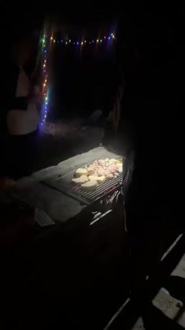 A man and woman stand near a grill with food on it. There are lights strung above them. The video then shifts to a dark scene with red lights in the distance.