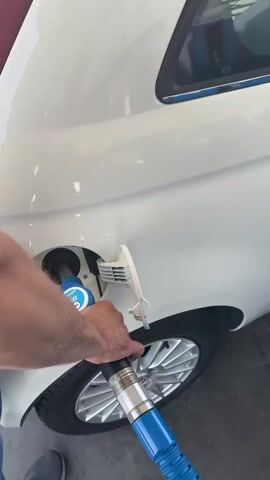 The video shows a person at a gas station filling a white car with gasoline. The camera focuses on the car's gas tank and the gas pump nozzle. The nozzle is blue and has a white and blue logo. The gas pump display shows the amount of fuel dispensed, the cost, and the price per liter. The price per liter is 1.903 euros. The video also shows the car's wheel and a part of the gas station's pump. The car appears to be a small hatchback. The environment is a typical gas station with blue pumps and a white digital display. The lighting suggests it is daytime. The video is shot from a slightly high angle, looking down at the car and the pump.