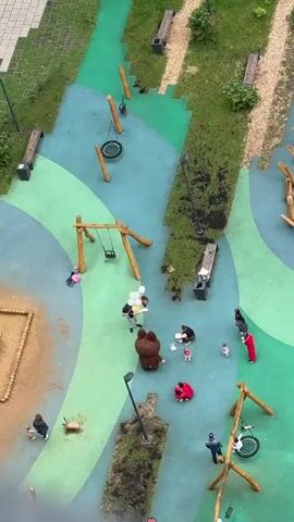 An aerial view captures a playground with several people and objects. In the center, a person in a brown bear costume stands near another person holding a cluster of balloons. A woman is seated on the ground with a child, and another child in a pink outfit walks nearby. Further to the right, two people in red and pink outfits are interacting, and a person in a red outfit is also present. To the left, a child is near a swing set. A bicycle is propped against a wooden structure on the right side of the frame. Benches and lampposts are also visible throughout the playground.