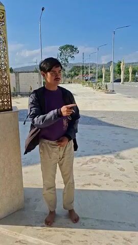 A young person walks barefoot on a paved outdoor area. They are wearing a purple t-shirt, tan pants, and a dark jacket. The person walks from left to right, then turns to walk away from the camera. The background shows a blue sky, some trees, white posts, and a golden structure.