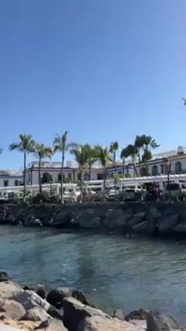 Certainly! Here's a detailed description of the scene:

**Scene:** The video depicts a vibrant beach scene on a sunny day.

**Environment:**
*   The environment transitions from a rocky bank near a canal or waterway, to a wooden boardwalk, and then to a sandy beach.
*   Palm trees and white buildings with dark trim are visible along the waterway.
*   The beach is packed with beachgoers, primarily sunbathers.
*   A large rocky hill is visible in the background, with buildings constructed on it as well.

**People:**
*   Individuals of various ages and ethnicities are seen on the beach.
*   Many are sunbathing on lounge chairs.
*   Some people are walking along the boardwalk and beach.
*   A shirtless man with sunglasses on his head walks by the camera.
*   Other individuals are dressed in typical beach attire (swimsuits, shorts, t-shirts).

**Objects:**
*   Lounge chairs are arranged in dense rows across the beach.
*   Blue and white striped towels cover some of the lounge chairs.
*   A kiosk with red and blue flags stands near the beach.

**Actions & Interactions:**
*   People are relaxing, sunbathing, walking, and generally enjoying the beach.
*   The camera pans across the scene, providing a broad view of the beach and surrounding area.
*   People walk along a boardwalk.
*   The water is calm and visible beyond the beach area.

**Overall Impression:** The scene evokes a lively and popular tourist destination on a warm, sunny day.