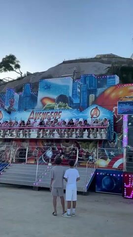 The media file captures a scene at an amusement park in Canet de Mar, Spain, likely during the late afternoon or early evening, as indicated by the soft, warm light and clear sky. The primary focus is a vibrant amusement ride themed with Marvel's Avengers characters. A large mural adorns the ride's backdrop, featuring prominent Avengers like Hulk, Iron Man, Captain America, and Vision, set against a backdrop of a stylized cityscape and a fiery, exploding planet. The "AVENGERS" logo is clearly visible across the mural.

The ride itself consists of a raised platform with rows of seats, each equipped with safety restraints. Several people, predominantly young individuals, are seated on the ride, with many appearing to be enjoying themselves, raising their hands and looking excited. The ride is decorated with numerous colorful lights, some of which are illuminated.

In the foreground, two young individuals, likely teenagers, stand facing the ride, observing it. They are casually dressed, one in a beige t-shirt and shorts, the other in a white t-shirt and white shorts. The ground beneath them is a sandy or dusty surface, typical of an outdoor amusement area. To the right of the ride, a tall pole with lights and a speaker can be seen, suggesting the presence of further park infrastructure. The overall atmosphere appears to be one of fun and entertainment, with the exciting theme of the ride and the visible enjoyment of the riders contributing to a lively, festive mood. The background shows a hillside with some vegetation and buildings, placing the scene within a coastal town.