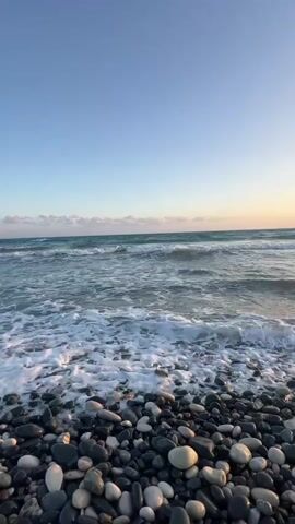 The video shows a view of the ocean from a pebble beach. The sky is clear and blue with a few clouds on the horizon. The waves are crashing onto the shore, and the water is a clear blue. The beach is covered in smooth, round pebbles of various colors, including black, gray, and white. There are no people visible in the foreground or mid-ground, but a few small figures can be seen swimming in the distance on the left side of the frame. The lighting suggests it is either early morning or late afternoon.
