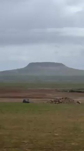 Here's a description of the scene:

The video shows a wide, flat landscape under an overcast sky. In the distance, a flat-topped hill or mesa is visible. The foreground is a grassy field, with a section of brown earth in the midground. A small pile of what appears to be rocks is to the right of the frame. The overall impression is one of a vast, open space with a distinct geological feature in the distance.