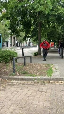The video captures a person in a red hoodie and baggy jeans performing a stunt on an electric scooter in an urban setting. The individual rides the scooter towards a metal barrier, jumps onto the barrier with the scooter, balances momentarily, and then successfully rolls off the barrier onto the sidewalk. The action occurs on a paved surface near a grassy area with trees and shrubs. Other people are visible in the background riding similar scooters or walking along the sidewalk. A building with a parking sign is also visible.