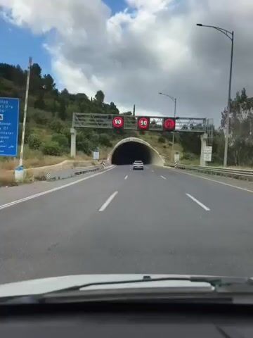 The video shows a car driving into and through a highway tunnel. The car approaches the tunnel entrance, passing under a sign indicating a speed limit of 90. The tunnel entrance is a wide concrete arch with "5.0" printed on it. Inside, the tunnel is well-lit with overhead lamps. The car follows lane markings, passing other vehicles. Ventilation equipment is visible on the ceiling.