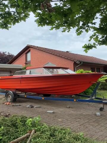 The video shows an orange boat on a blue trailer in a residential setting. The boat is a cabin cruiser style with many windows and is placed on a trailer parked on a paved driveway or area. Behind the boat is a brick house with a red tile roof and a green lawn. The sky is overcast and trees are visible in the foreground and background, providing a slightly obscured view of the scene. The camera seems to be static, capturing a medium shot of the boat and its surroundings. The scene appears to be a personal property or a yard space.