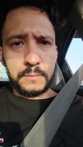 A medium close-up video captures a man seated in what appears to be the driver's seat of a car, wearing a seatbelt diagonally across his chest. He has dark, curly hair and a beard with a mustache, and is wearing a black t-shirt. A black earbud is visible in his left ear. Throughout the video, his expression remains neutral to slightly pensive, and he looks directly into the camera.

The background visible through the car window suggests bright daylight with a clear blue sky, indicating it is daytime. An elevated structure, possibly a bridge or overpass, and distant buildings can be seen outside the window, confirming an urban or suburban setting. The location is Rímac, Peru. The man is not actively doing anything other than sitting in the car, and there is no visible text.