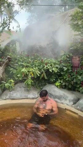 A man with tattoos is sitting waist-deep in a circular hot spring, surrounded by lush greenery and rocks. Steam rises from the rock face behind him. The water in the hot spring is a murky brown color.