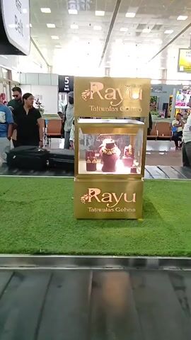 The video shows a jewelry display case next to a baggage carousel. The display case is gold-colored and features the name "Rayu Tatiwalas Gehna" in a large, stylized font. Inside the case, jewelry is displayed on purple stands. The jewelry appears to be ornate and gold in color. People can be seen in the background waiting for their luggage. The floor around the display case is covered in artificial green grass. The baggage carousel is silver and shows luggage moving along its conveyor belt.
