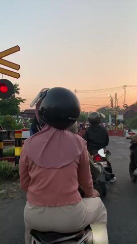 A low-angle vertical shot shows a group of people on motorcycles waiting at a railway crossing at sunset. In the foreground, a person in a pink top and beige pants sits on a black motorcycle, facing away from the camera. Several other individuals are visible in the background, also on motorcycles, waiting at the crossing. The sky is a gradient of orange and yellow, indicating dusk. A railway crossing barrier is partially visible on the left, and power lines crisscross the sky. The scene is static, with no movement visible from the people or vehicles.