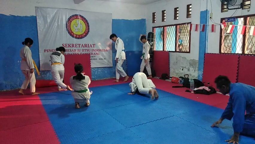 A group of people, primarily young individuals wearing white martial arts gis, are practicing Jiu-Jitsu on a blue and red mat in an indoor training area. The scene is likely a dojo or a similar facility.

The subjects are engaged in various movements, suggesting a training session or sparring. In the foreground, a person in a white gi is on their back with their legs in the air, possibly demonstrating or practicing a technique. To their right, another individual in a white gi is sitting and observing. Further back, two people are kneeling on the mat, one appearing to be in a defensive position while the other is on their hands and knees. To the left, two individuals in gis are standing, one with a yellow belt, and another in a white gi.

Visible objects include a punching bag, a kettlebell, a water bottle, and bags, all placed near the edge of the mat. The walls are painted with blue and red, and there are windows with blinds in the background. A large banner with text and a circular emblem is displayed on the wall behind the practitioners.

The lighting suggests it's daytime, possibly late morning or afternoon. The overall atmosphere appears to be focused and active, with participants engaged in physical training.

Visible text on the banner reads: "SEKRETARIAT PENGURUS BESAR JI JITSU INDONESIA KABUPATEN TANGERANG" and below that, "PERUM MU... MUNUL - SOLE...". Indonesian flags are also visible in the background, indicating the location is likely Indonesia, further supported by the text on the banner which mentions "Kabupaten Tangerang".