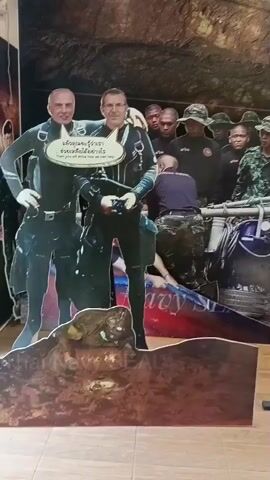 The video pans across a series of life-sized cardboard cutouts of people standing in front of a large, printed backdrop that depicts a cave interior with several figures in the background that appear to be divers or rescue personnel. The cutouts are arranged in a line and display various individuals in different attire. The first cutout on the left shows two men in diving suits. The next cutout features a person in a white shirt, a yellow scarf, and a blue cap. Following this are cutouts of individuals in military camouflage uniforms, one in a dark uniform with a black helmet, and another in a black shirt and jeans. The backdrop behind the cutouts shows a dimly lit cave environment with people in the background, some wearing helmets and tactical gear, and equipment that suggests a rescue or exploration operation. The floor in the foreground has black and yellow striped hazard tape.