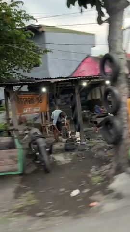 The video shows an outdoor scene, likely a roadside workshop, at dusk. In the foreground, a person is working on what appears to be a motorcycle engine. They are crouched down, wearing a light-colored shirt and dark pants. Several tires are visible, some stacked against a tree on the right side of the frame, and others near the workshop area. The workshop itself is a simple structure with a corrugated metal roof, open on one side, revealing workbenches and tools. A sign with orange lettering is mounted on the workshop wall, partially obscured but readable as "REGION 16 KRETEK". A motorcycle is parked to the left of the person. The ground is dirt and appears wet. In the background, buildings and trees are visible. The overall impression is of a humble, functional workspace.