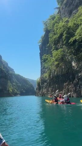 The video captures a group of people kayaking on a clear, turquoise lake, surrounded by towering, tree-covered cliffs. The sun is shining brightly, indicating a warm, clear day. The kayakers are in various colorful kayaks, mostly yellow and red, and appear to be enjoying a leisurely excursion. They are wearing life jackets for safety. The sheer scale of the cliffs suggests a canyon or a large gorge, characteristic of the mountainous region of Bezuje, Montenegro. The water is calm, reflecting the blue sky and the lush greenery of the cliffs. The overall mood is one of peace and outdoor adventure. There is no visible text in the media file.
