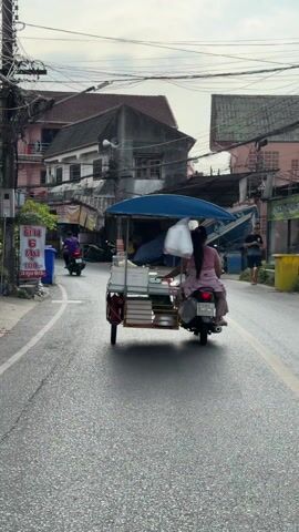 A woman driving a motorcycle with a cart attached moves down a street lined with buildings on both sides in an urban setting. The cart is covered by a blue umbrella and is filled with white containers. The woman is wearing a pink dress and has a white bag hanging from her head and neck. Other motorbikes pass in the opposite direction, and a few pedestrians are visible on the sidewalk. The day is overcast, and the pavement is wet. Power lines crisscross above the street. A sign in Thai is visible on the left.