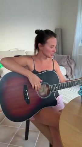 A woman sits on a chair holding an acoustic guitar, looking at the camera. She is wearing a black tank top and pink shorts. She smiles and raises her right hand as if to wave. A table with a blue mug is visible to her right. The background shows a couch and a window.