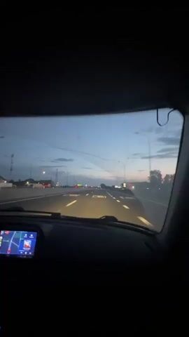 The video is filmed from inside a car at dusk or dawn. The camera faces forward, showing the car's dashboard and windshield. On the dashboard, a GPS navigation system is visible on the right, displaying a map. To the left, a car's instrument cluster shows various gauges and indicators. Below the navigation screen, a car stereo with illuminated blue buttons is present. The windshield shows some streaks or cracks. Outside, a highway stretches ahead, with other vehicles visible in the distance. Streetlights and overhead power lines line the sides of the road. The sky is a dusky blue with some clouds. The overall scene depicts a car traveling on a highway as daylight fades or begins to appear.